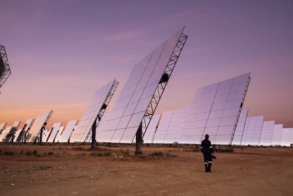 Ein riesiges Solarkraftwerk bei Sonnenuntergang oder Sonnenaufgang, mit Reihen großer, reflektierender Solarmodule, die zum Himmel ausgerichtet sind. Eine einzelne Arbeiterfigur geht im Vordergrund und hebt die Größe der erneuerbaren Energieanlage hervor. 