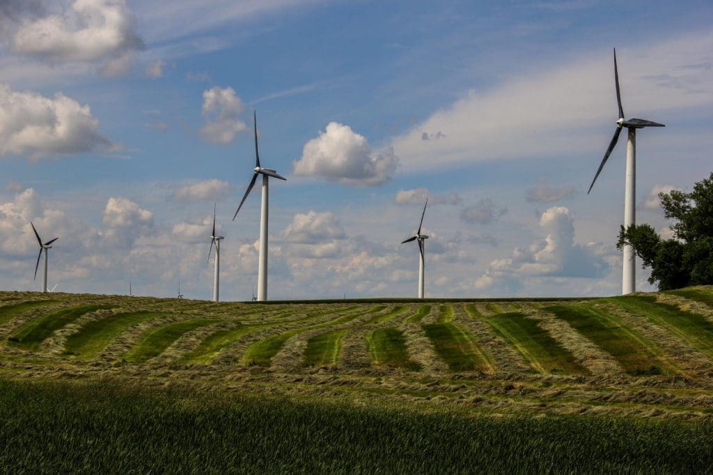 Mehrere große weiße Windkraftanlagen stehen auf einem hügeligen grünen und braunen Feld unter einem bewölkten blauen Himmel.