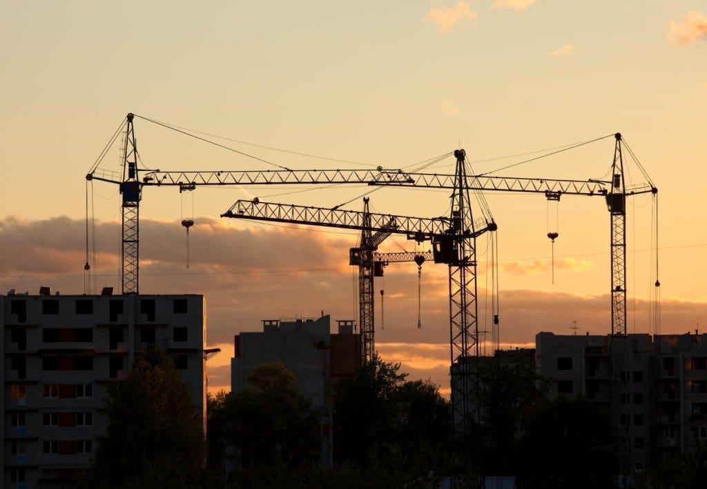 Silhouetted tower cranes dominate the skyline over buildings during a golden, cloudy sunset.