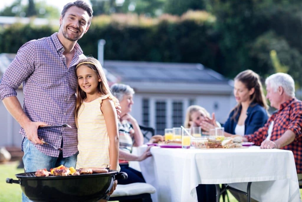 Lächelnder Vater und Tochter stehen am Grill, während die Familie im sonnigen Garten im Freien speist.
