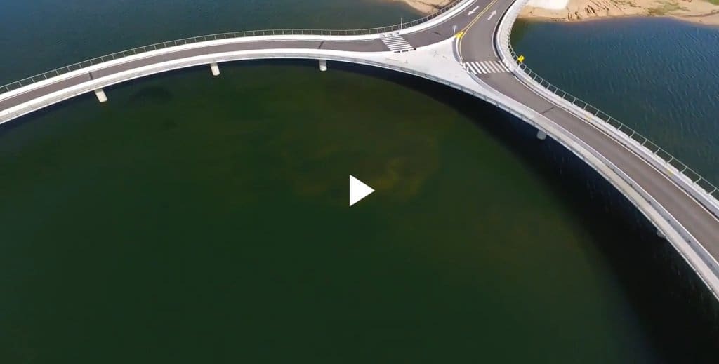Aerial view of a modern curving bridge structure spanning dark green water near a rocky shore.