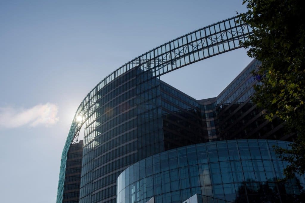 Tiefwinkelaufnahme eines geschwungenen Glaswolkenkratzers mit einer markanten Überführungskonstruktion vor einem strahlend blauen Himmel.